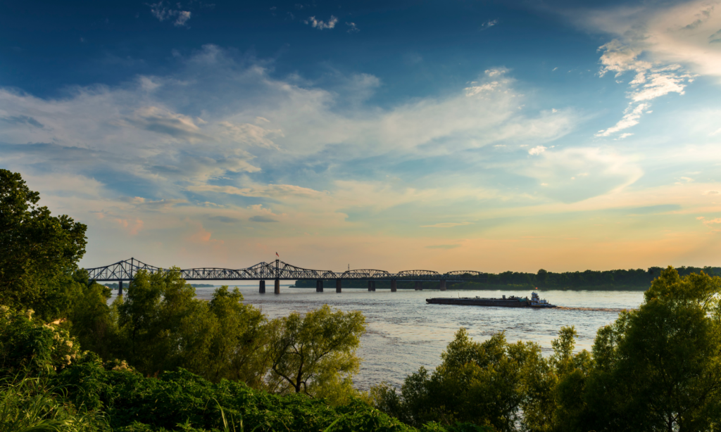 Sunset over the Mississippi River in Vicksburg, Mississippi, with a towboat passing beneath a historic bridge.
