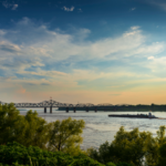 Sunset over the Mississippi River in Vicksburg, Mississippi, with a towboat passing beneath a historic bridge.